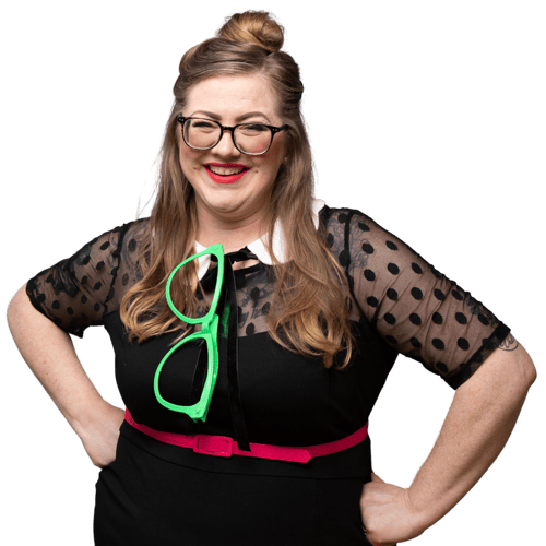 Maggie is shown wearing a black secondhand dress with Swiss dot yoke, a hot pink belt, and a removable collar made from fabric and ribbon scraps. She has an oversized pair of green glasses tucked into the neckline. She is smiling.